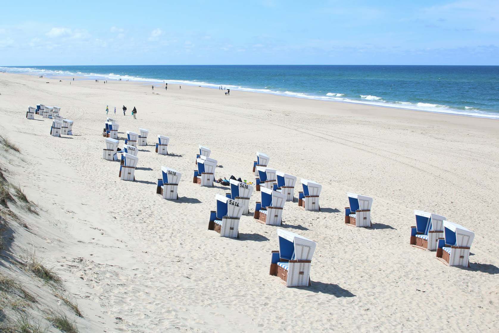 Sonniger Nordseestrand, mehrere Strandkörbe, im Hintergrund Spatzierende und Badegäste