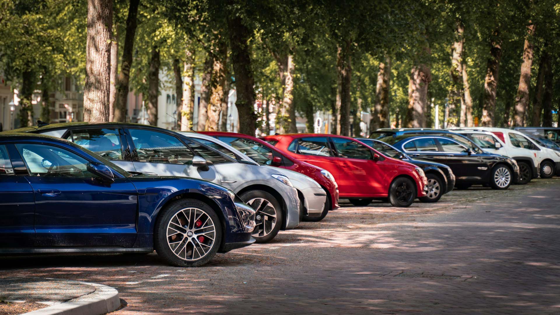 Autos parken nebeneinander am Straßenrand, dahinter Bäume und Mehrfamilienwohnhäuser