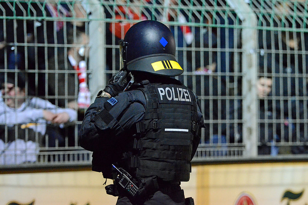 Schwer gepanzerter Polizeibeamter in Einsatzkleidung steht mit dem Rücken zur Kamera vor einem Stadionzaun, hinter dem Fußballfans zu sehen sind.