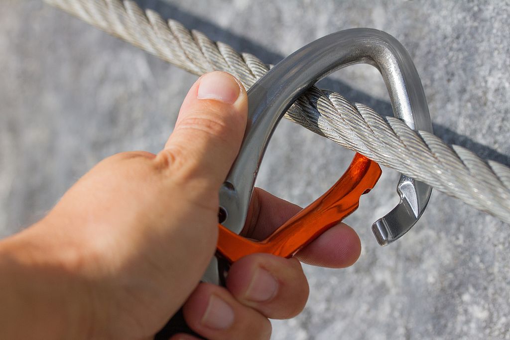 Wie Selbstständige sich absichern sollten. Hand hakt Karabiner in ein Drahtseil ein – Symbol für Sicherung und Stabilität beim Klettern oder Bergsteigen."