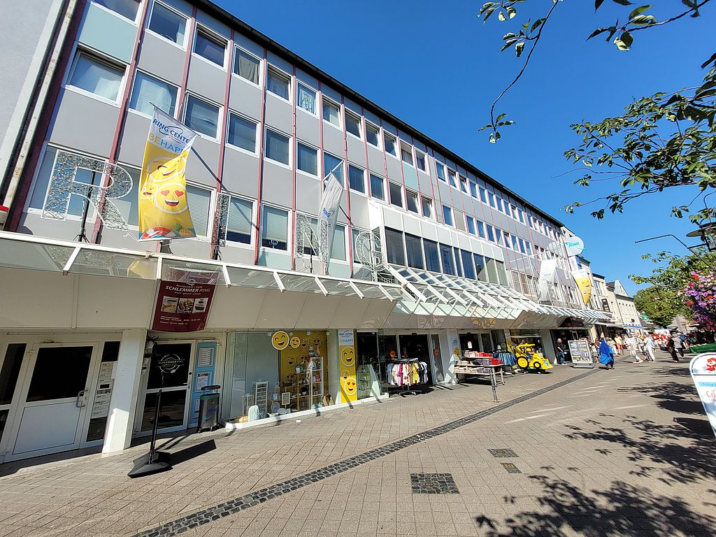 Die Stadt Dormagen kauft das Ringcenter in der Innenstadt. Mehrstöckiges Geschäftsgebäude 'Ringcenter' mit Schaufenstern, Flaggen und Passanten bei sonnigem Wetter in Dormagens Innenstadt.