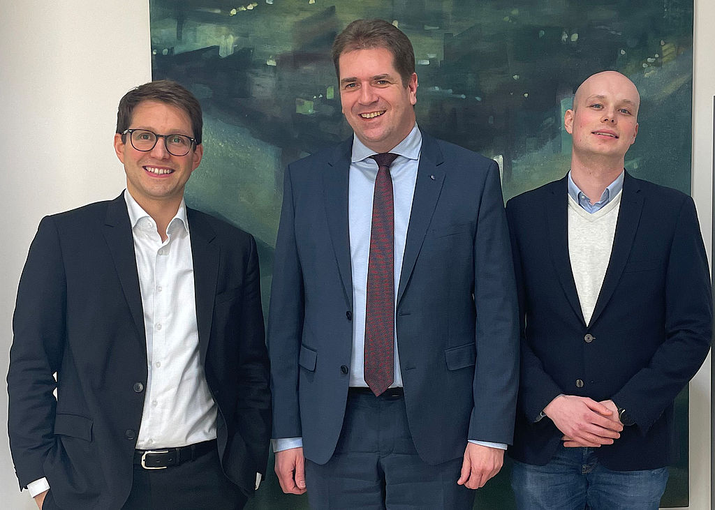 Der Finanzdezernent des Nordrhein-westfälischen und des Deutschen Städtetags Dr. Dominique Köppen (l.), der Vorsitzende des BdSt NRW, Rik Steinheuer, und BdSt-Haushaltsreferent Philipp Sprengel (r.) lächelnd vor einem angenehmen, abstrakten Bild, das mutmaßlich die Maße 2Meter mal Meter hat und in dunklem Blau sowie hellerem Grün gehalten ist. Die Farben sind spachtelartig aufgetragen.